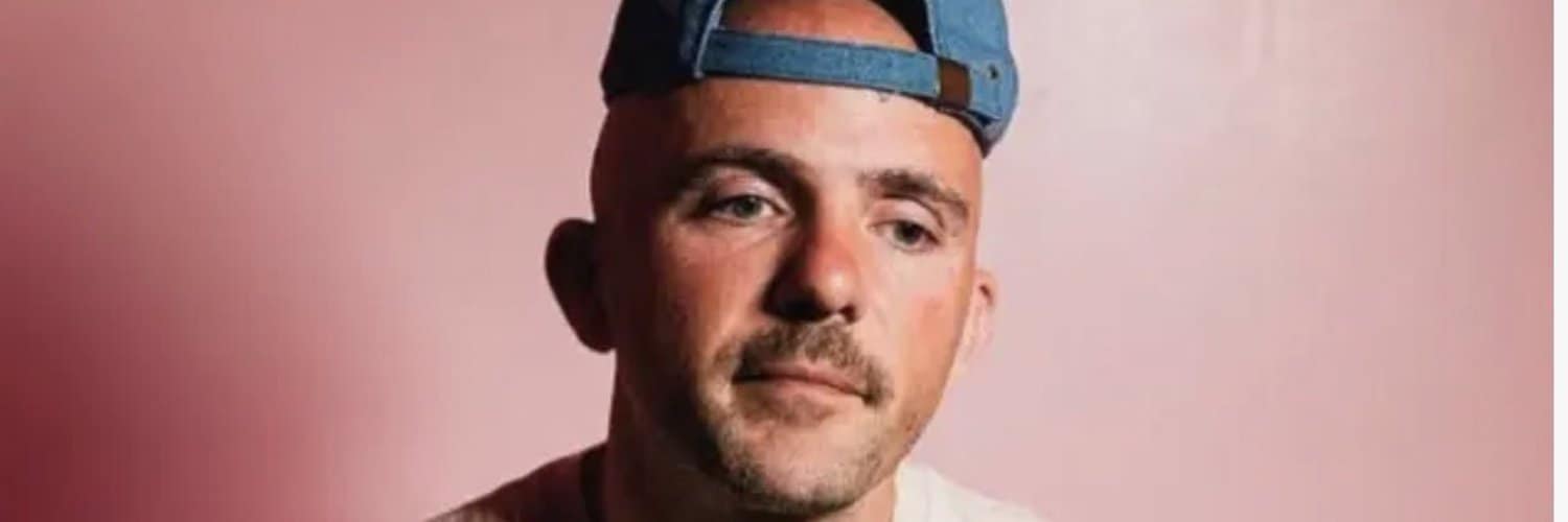 A young man with a serious expression wearing a blue cap backward, against a soft pink background, showcasing modern portrait photography.