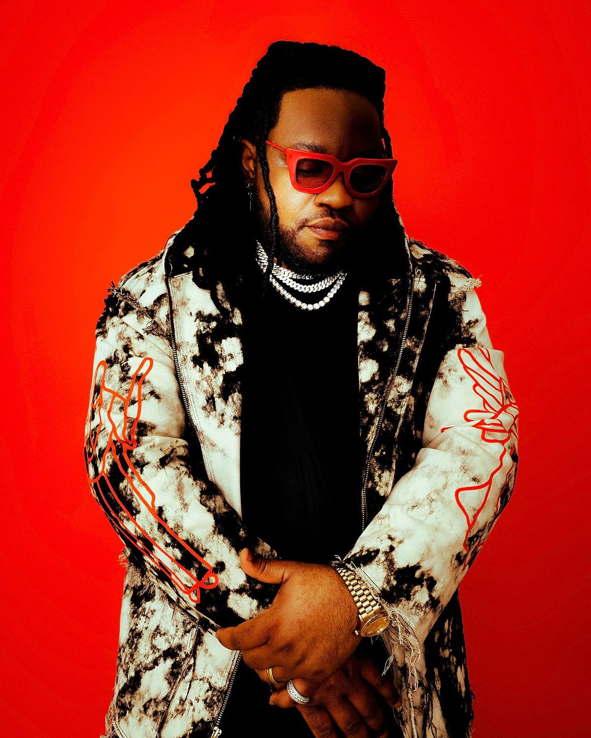 a photo of a man - Duncan Daniels wearing a patterned jacket, black shirt and red glasses, looking down while crossing his hands