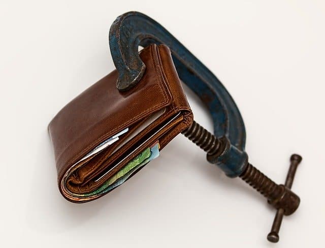 Overturned brown leather wallet being clamped by a large blue industrial clamp, symbolizing financial pressure or economic strain.
