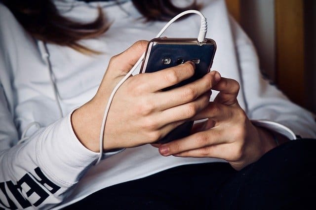 Young woman holding smartphone with earbuds, close-up shot, casual lifestyle, modern technology, mobile device usage, digital communication, attention to detail, urban setting, tech-savvy, everyday life.