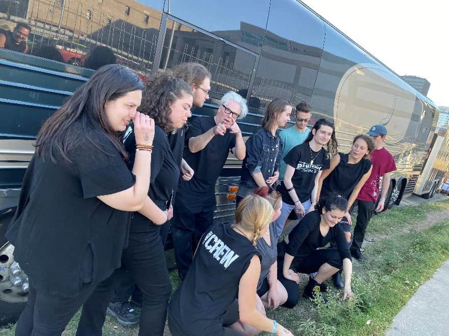 A diverse group of young people and an older woman gather outdoors beside a large black bus, engaging in a lively discussion or activity on a sunny day.