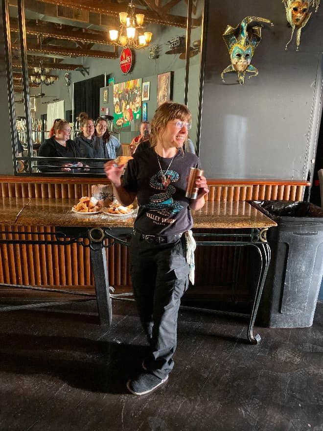 Cracked-open pizza on a wooden table at a lively restaurant with patrons in the background, festive masks hanging on the wall, and warm ambient lighting.