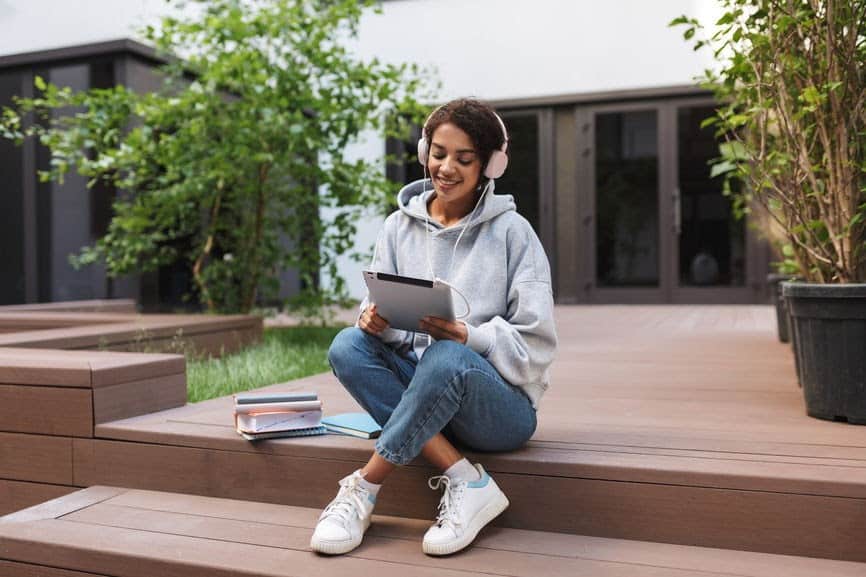 Relaxed young woman sitting outdoors on wooden steps, using a tablet with headphones, studying or working, surrounded by greenery, casual outfit, modern residential background.