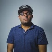 Man wearing glasses and a baseball cap, casual blue polo shirt, standing against a plain gray background.