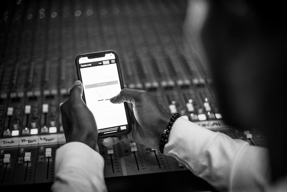 Music producer using smartphone for sound mixing and music technology at stereo console in studio, focusing on digital music production, audio engineering, music streaming, and tech innovations.