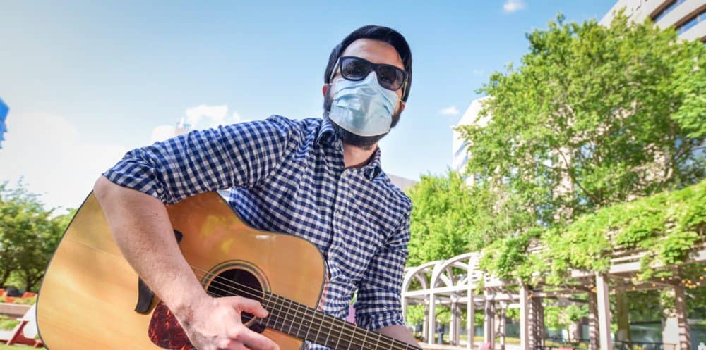 Percussionist, playing acoustic guitar outdoors, wearing sunglasses and face mask, vibrant park scene with green trees and blue sky, entertainment, live music, outdoor performance, social distancing, pandemic safety.