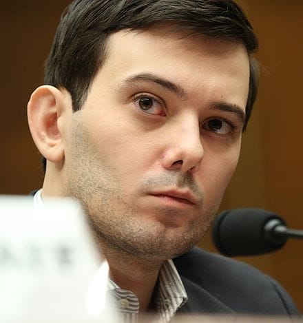 Caucasian man in suit speaking at a conference or panel discussion with microphone, focused and attentive.