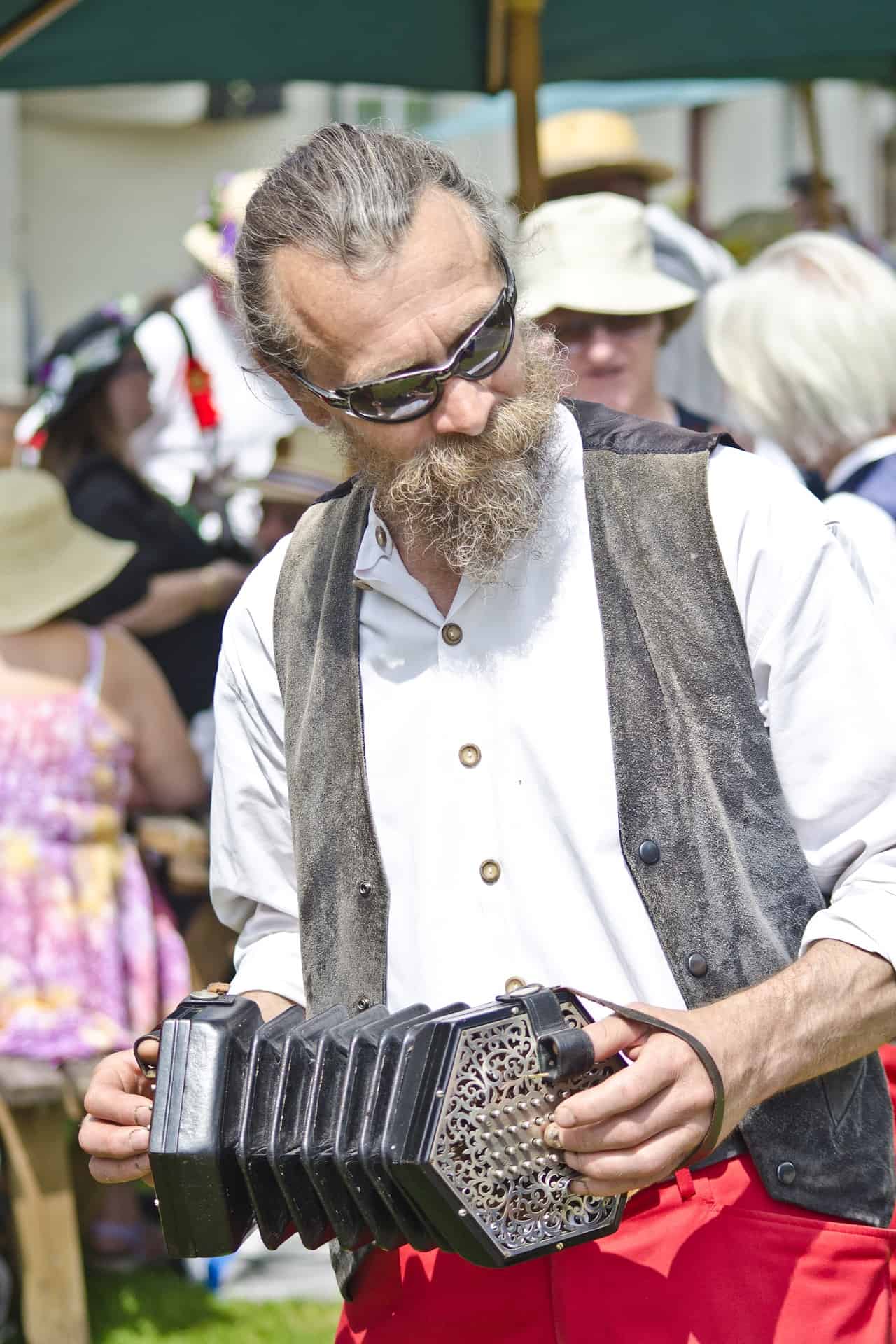 Vintage man playing concertina at outdoor festival, diverse crowd in background, casual summer attire, music event, folk music, summer festival atmosphere, outdoor entertainment, musician enjoying the moment.