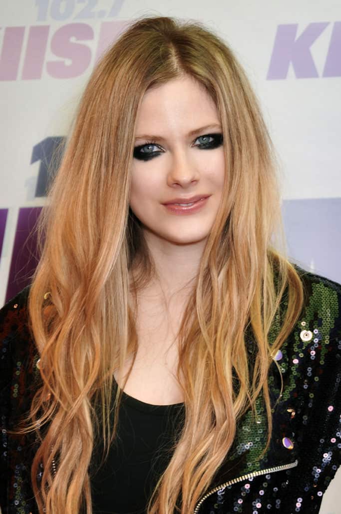 A young woman with long, wavy strawberry blonde hair, striking blue eyes, and bold makeup, wearing a black outfit with a sequin jacket at a media event or red carpet.