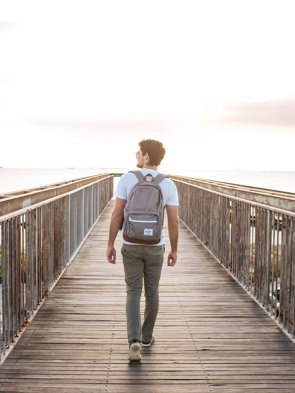 Young man with backpack walking on a wooden bridge towards the bright horizon, exploring nature and outdoor adventure, ecco-friendly lifestyle travel, relaxation, and scenic views.