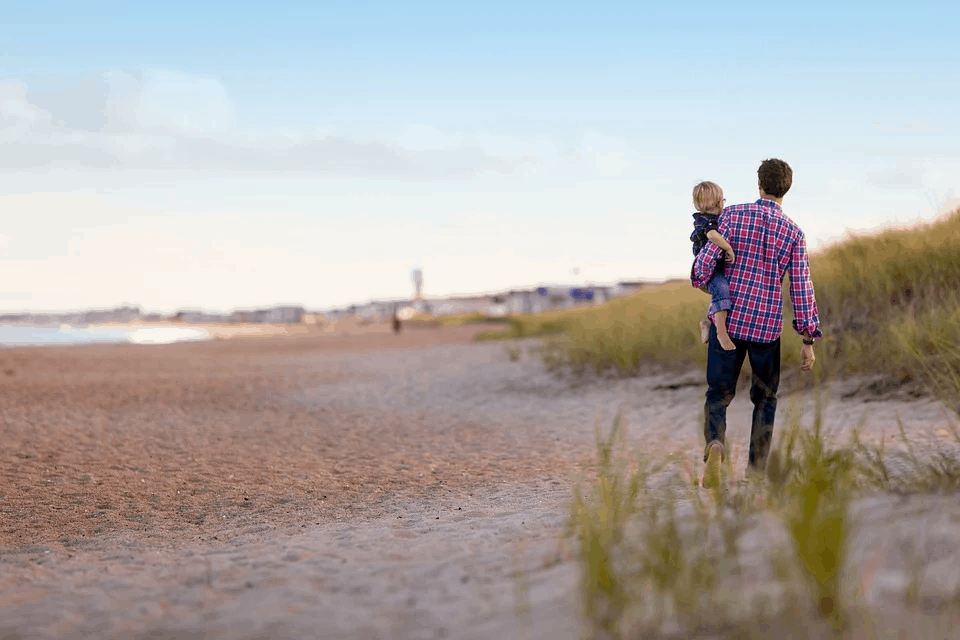 A man walking on the beach holding a child in his arms, enjoying a day outdoors, with sand dunes and a clear sky in the background.