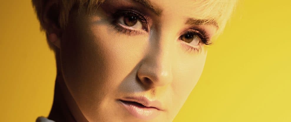 Highlights of a woman with short blonde hair and makeup, related to music and entertainment industry, vibrant yellow background, professional portrait, Hypebot featured image.