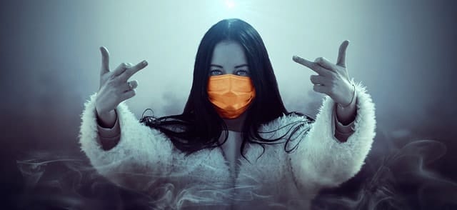 Slim woman wearing orange face mask making rock gesture with hands in foggy environment, modern urban style, dark hair, mysterious vibe, expressive pose, trendy fashion statement, artistic photography.