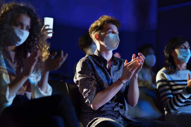 People wearing face masks attending a concert or event, clapping and enjoying entertainment in a dark indoor setting, emphasizing health safety measures at public gatherings.