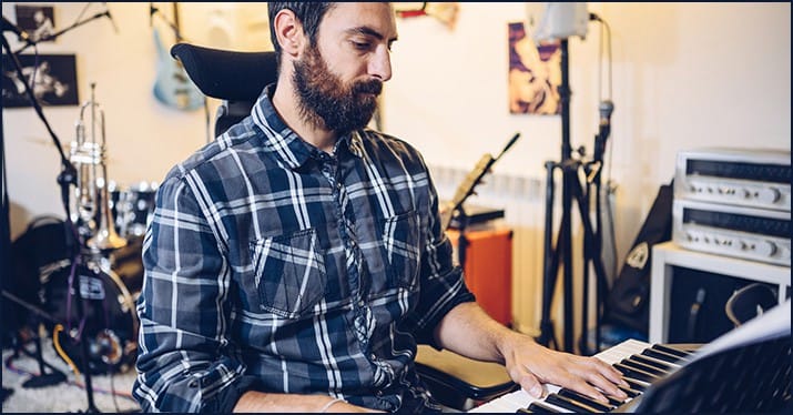 Music producer working at a keyboard in a home studio with musical instruments and equipment for music production and songwriting, Hypebot.