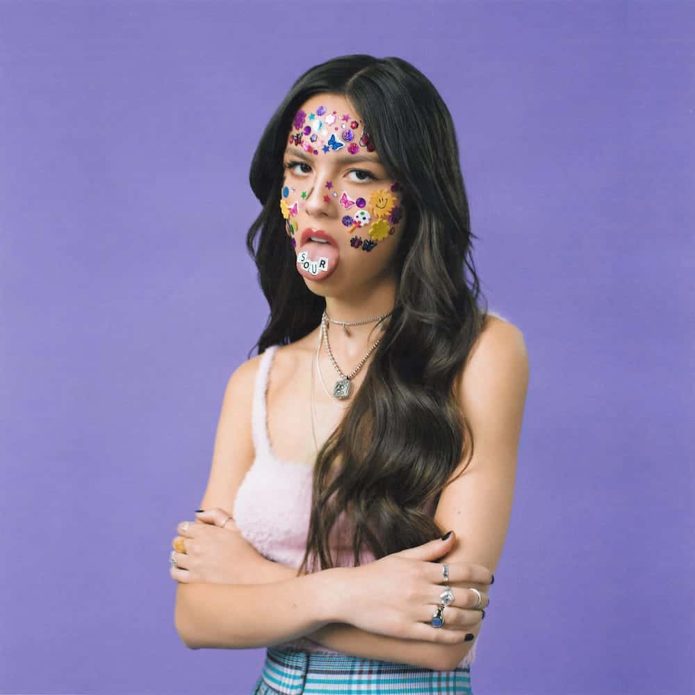 Colorful sticker-covered woman with playful expression, holding a "SOUR" candy, vibrant purple background, showcasing modern pop culture, youth fashion, and creative self-expression at Hypebot.