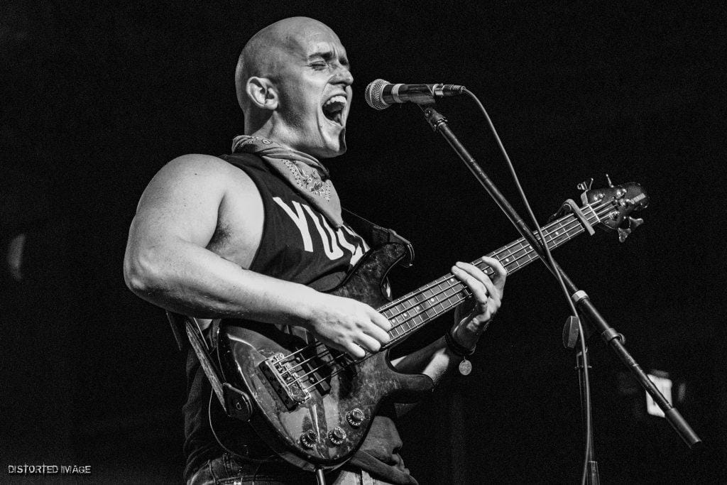 Bald musician passionately singing and playing electric bass guitar on stage at a concert, black and white image capturing energetic performance.