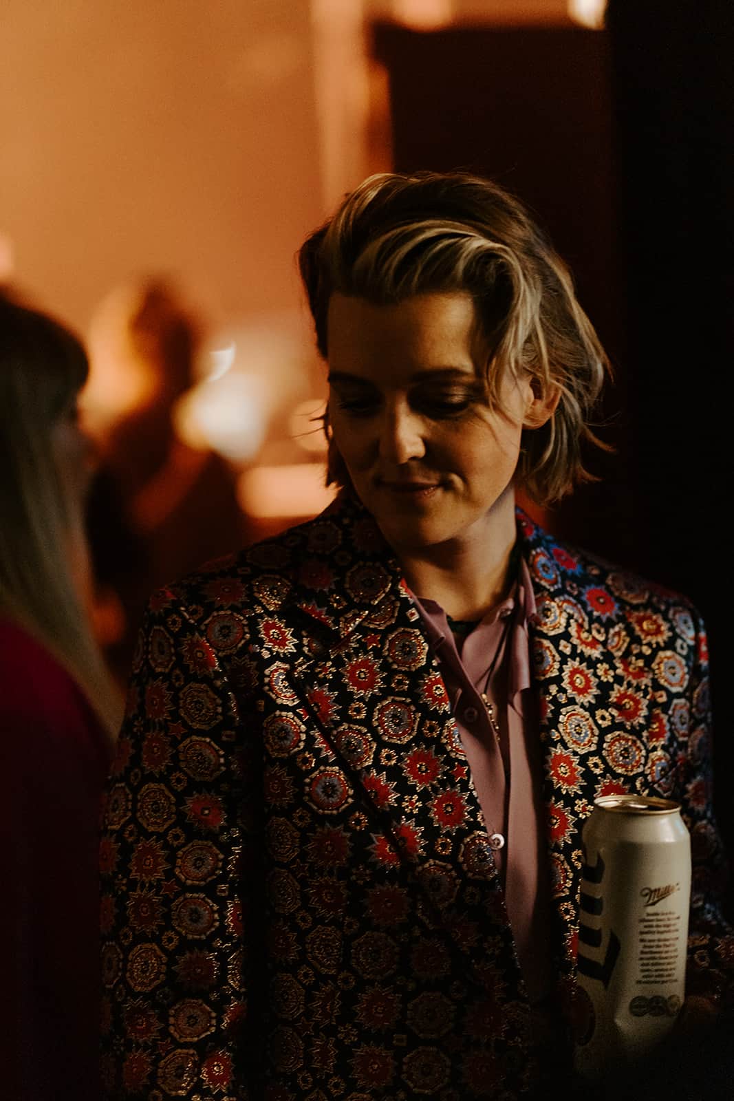 Stylish woman at a music event holding a drink, dressed in a vibrant floral blazer, in a lively nightlife atmosphere with warm lighting and blurred background.