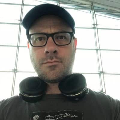 Close-up of a man wearing glasses and headphones, standing in an airport terminal with large windows, capturing the essence of travel, music, and modern technology.