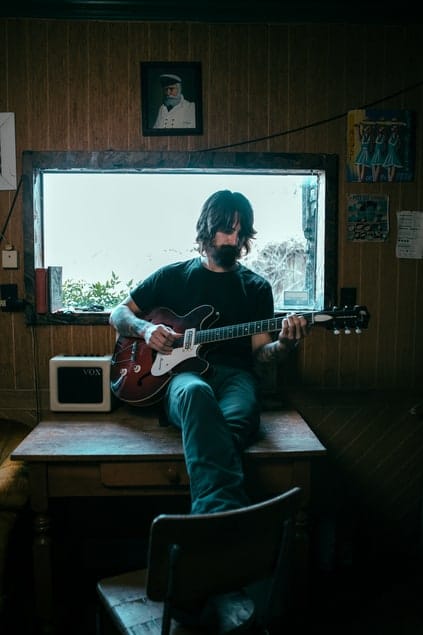 Man playing electric guitar in cozy room with wood-paneled walls and art posters, natural light from window, music studio atmosphere, music inspiration, indie musician, creative space, musical performance, artistic environment, Hypebot.