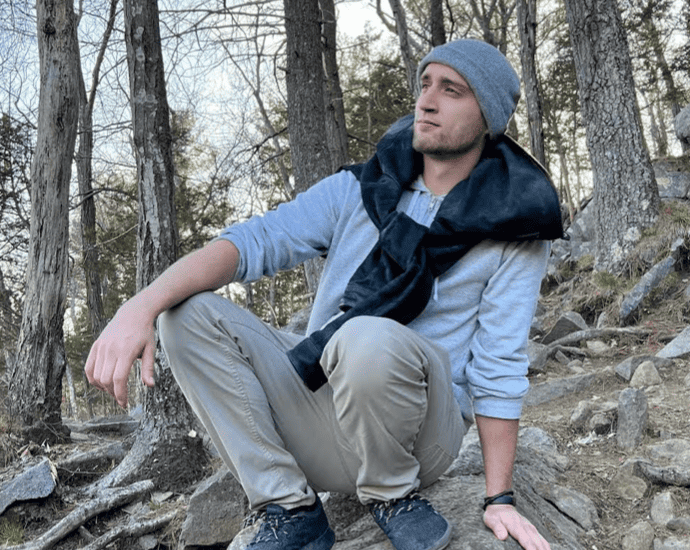 A young man sitting on rocky terrain surrounded by trees, dressed in outdoor clothing, looking contemplative in a natural forest setting.