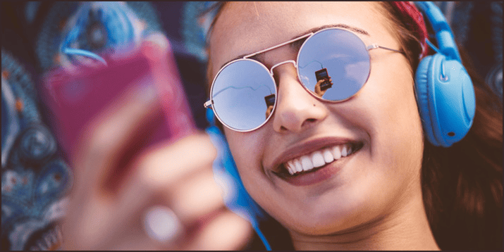 A young woman with sunglasses and headphones smiling while looking at her phone, enjoying music and digital entertainment at the beach.