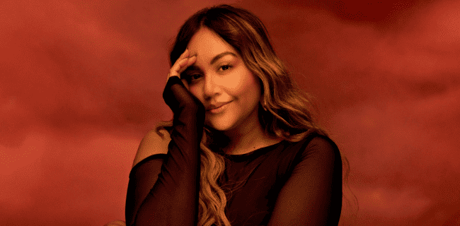Young woman in black sheer top smiling and posing for a marketing or branding photo with warm red background, suitable for music, entertainment, or digital media content.