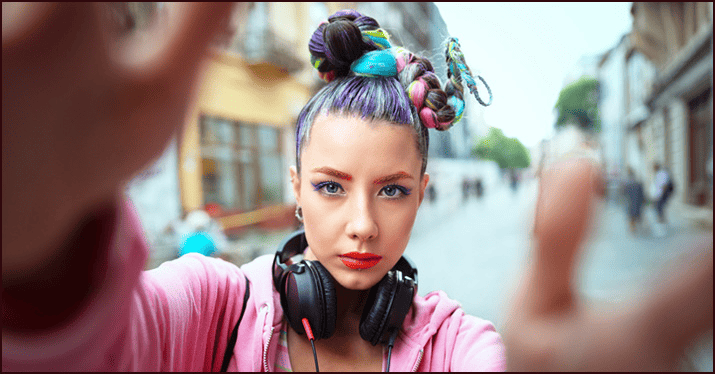 Young woman with colorful braided hair taking a selfie in an urban street scene, wearing headphones around her neck, showcasing modern youth culture and music fashion.