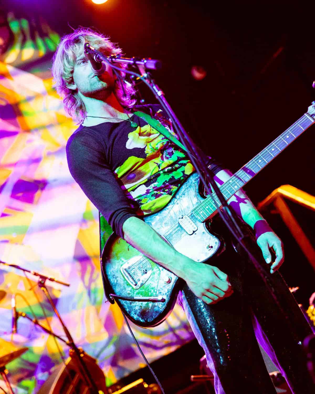 Vibrant rock concert scene featuring a guitarist with long blonde hair playing an electric guitar under colorful stage lighting. The backdrop displays psychedelic visuals, emphasizing live music, performance, and music industry themes for SEO.