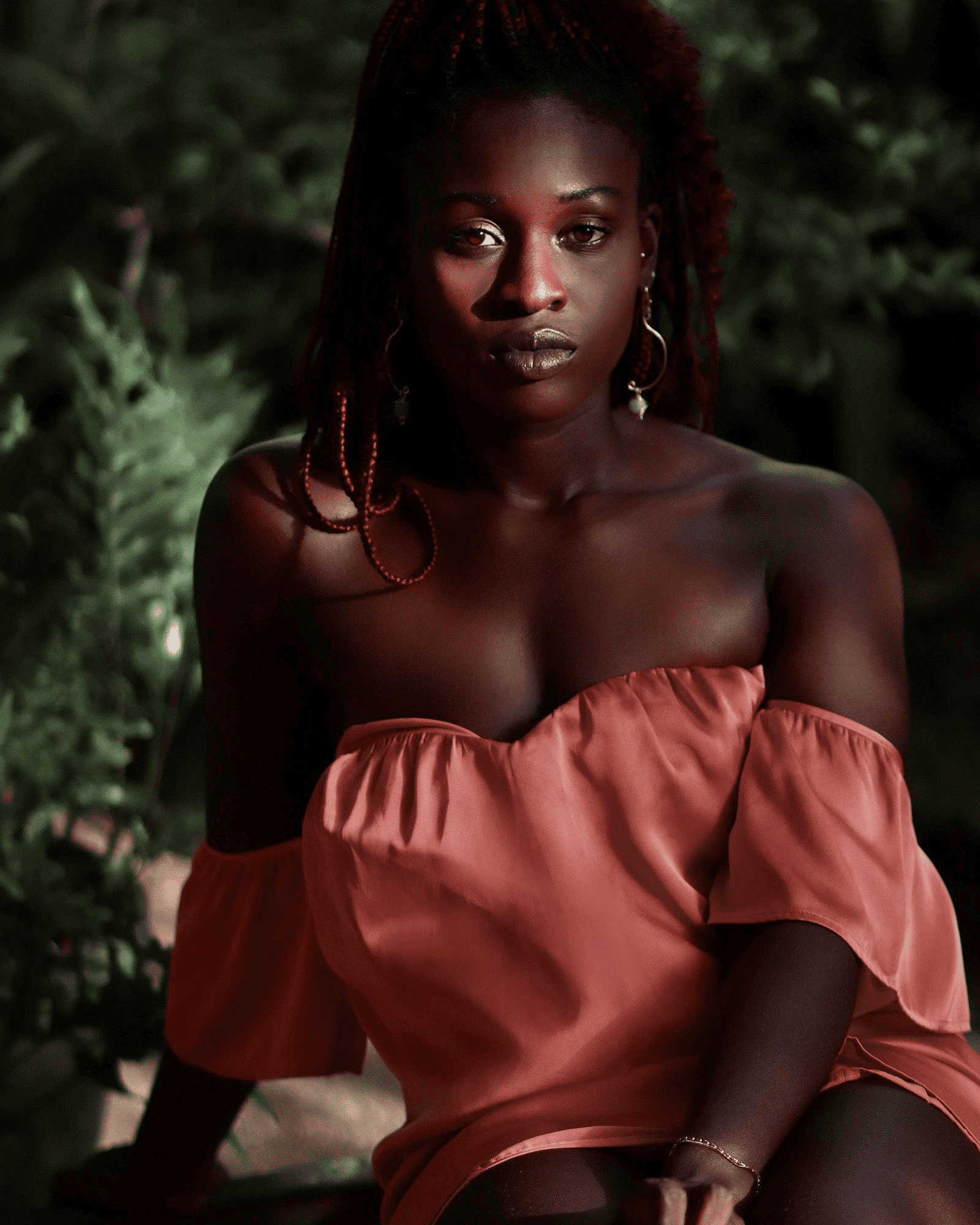 Gorgeous dark-skinned woman with natural makeup and red dreadlocks, wearing an off-shoulder peach dress and jewelry, posing outdoors in lush green garden for fashion or beauty photoshoot.