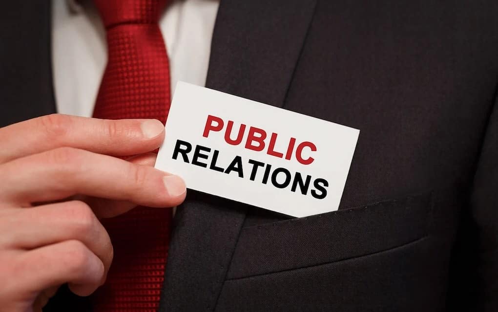 Corporate businessman holding "Public Relations" card in suit and red tie, emphasizing communication and media engagement skills.