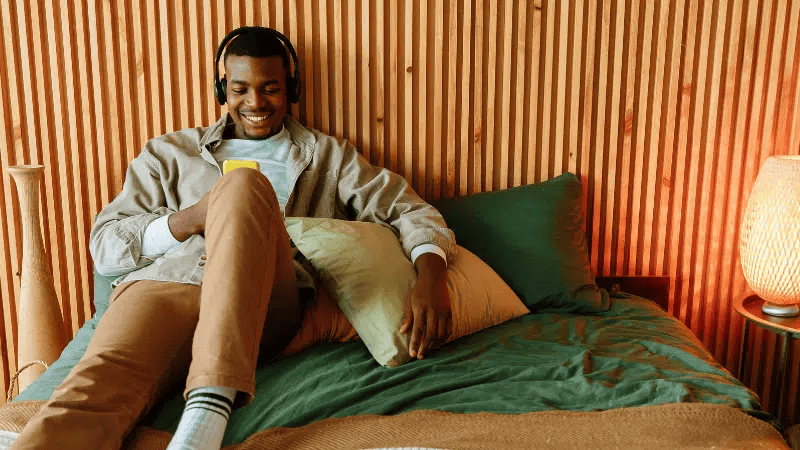 Young man enjoying music with headphones, smiling on a cozy bed in a modern bedroom.