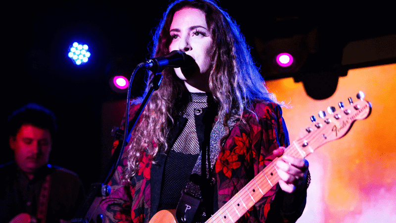Young female musician playing an electric guitar and singing into a microphone during a live performance at a music venue with vibrant stage lighting shooting blue, purple, and orange hues.
