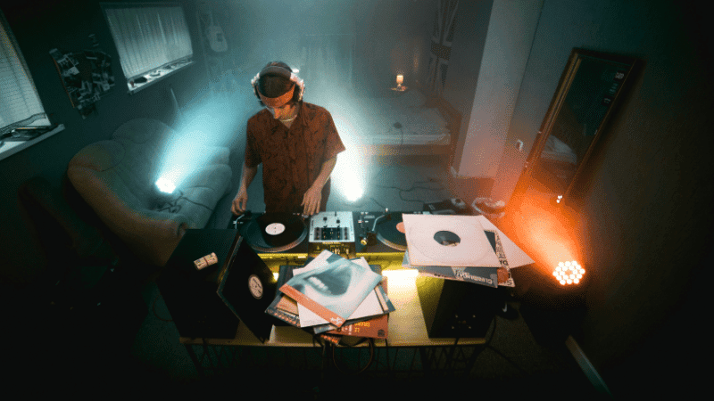DJ playing vinyl records and digital music on a mixer with vibrant lighting in a cozy, dimly lit room for a music event or party.