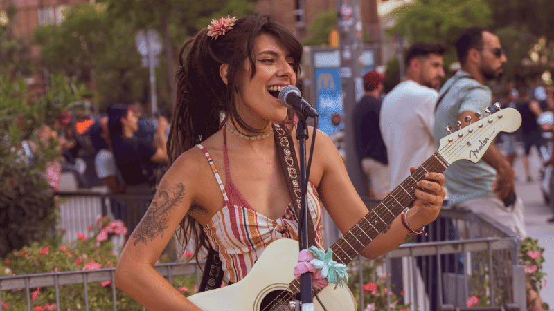 Young woman performing live music with guitar at outdoor festival, promoting music industry events, artist performances, and live entertainment coverage for Hypebot.