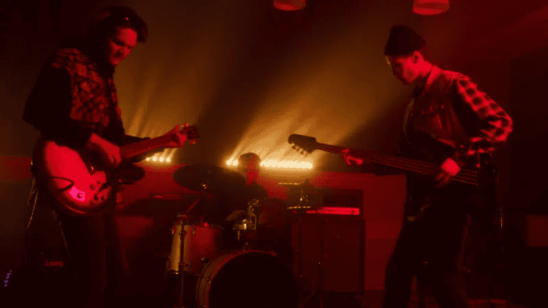 Guitarists and drummer performing live on stage in a dimly lit, vibrant red-tinged concert setting, capturing mid-performance energy and musical passion.