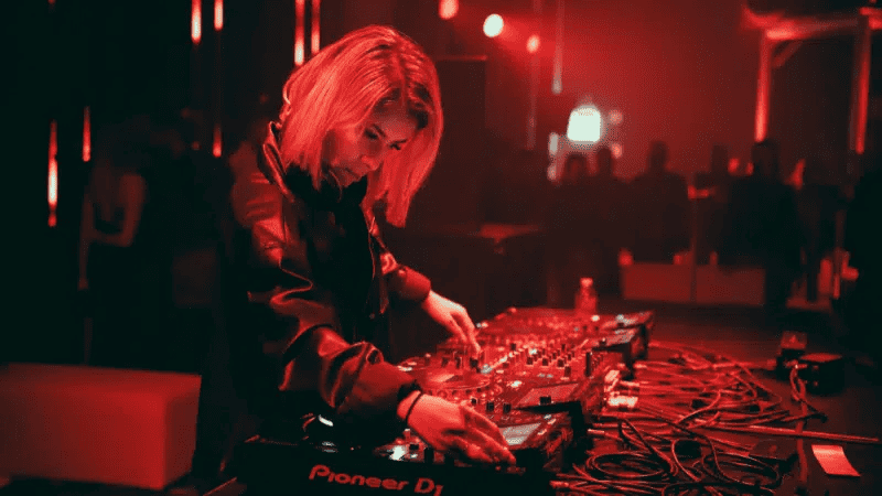 Mixing DJ, electronic music, and nightclub scene at Hypebot event; female DJ in a vibrant red-lit club setting using Pioneer DJ equipment for nightlife entertainment.