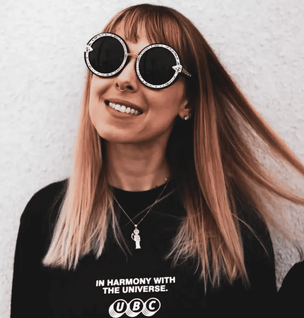 Breathtaking woman with stylish round sunglasses and ash blonde hair, wearing a black T-shirt with music-inspired text, standing against a white textured wall, embodying music culture and trendy fashion for Hypebot image.