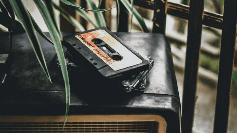 Cassette tape labeled "compact cassette" resting on a vintage audio device, surrounded by green plants and wooden furniture, representing retro music, audio nostalgia, and vintage audio equipment.