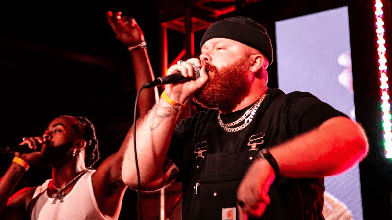 People performing live hip-hop music at a concert stage, artist wearing black beanie and chains, vibrant red and purple stage lighting, energetic music event, hip-hop culture and entertainment, Hypebot.