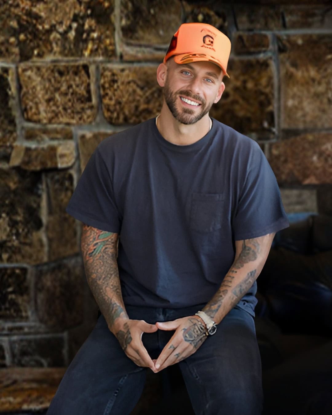 A smiling man with tattoos wearing a black t-shirt and orange cap, sitting against a stone wall, relaxed outdoor setting for lifestyle and music industry content.