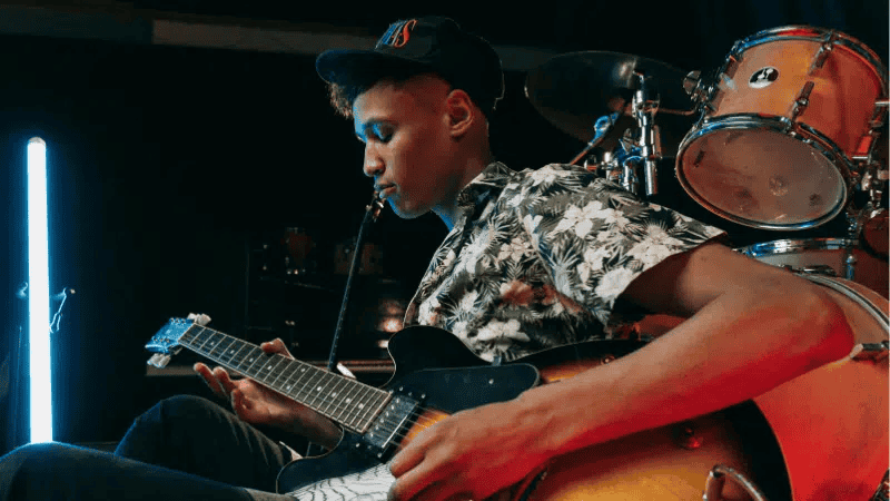 Young musician playing electric guitar in a modern music studio with drums in the background, showcasing music creation and performance.